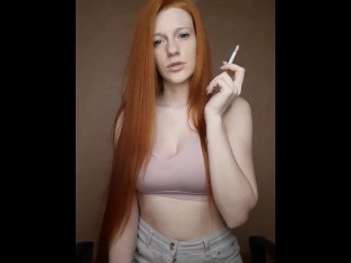 Рыжеволосая девушка с длинными волосами курит сигарету с коричневым фильтром.