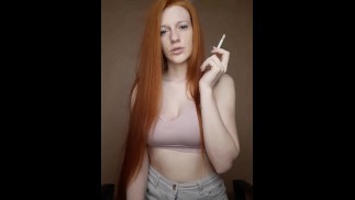 A red-haired girl with long hair smokes a cigarette with a brown filter.