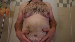 Primer plano del torso peludo obeso mientras está de pie desnudo en la bañera frotando o masajeando su vientre